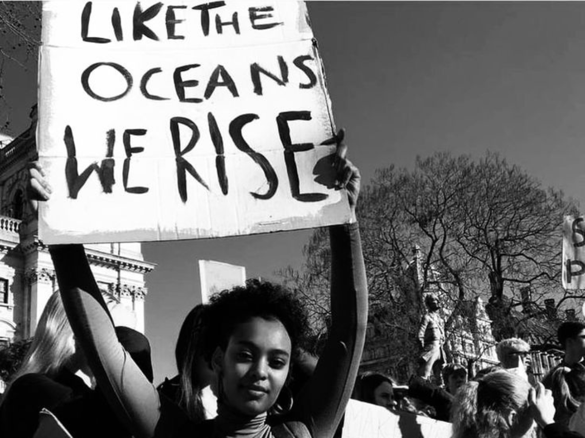 Black and white photo of a woman at a protest holding up a sign that says “Like the oceans we rise,” by Unknown Photographer
