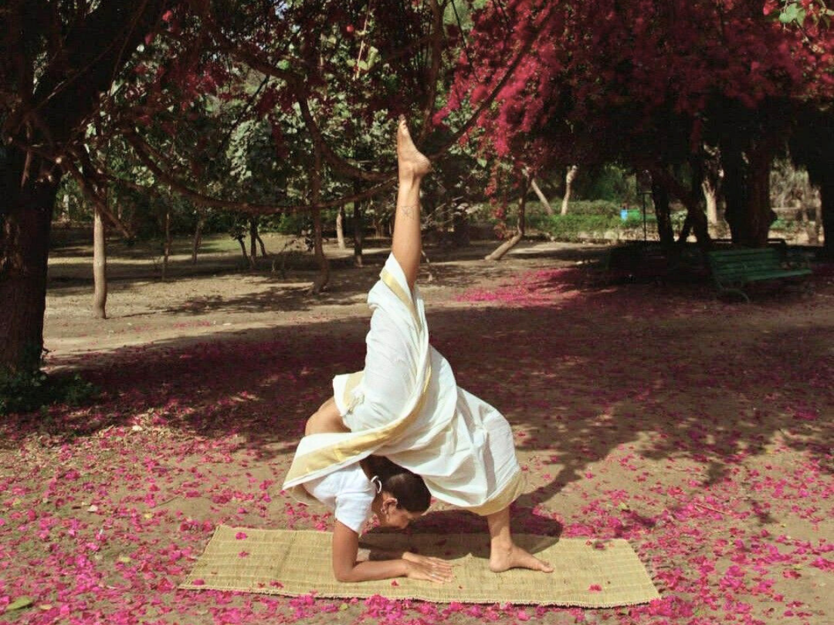 A South Asian yoga practitioner demonstrating an inverted backbending yoga asana (Eka Pada Viparita Dandasana) in a garden